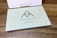 Custom 14pt uncoated matte postcards with light blue floral design and Ali Munn Jewellery logo on a wood grain background