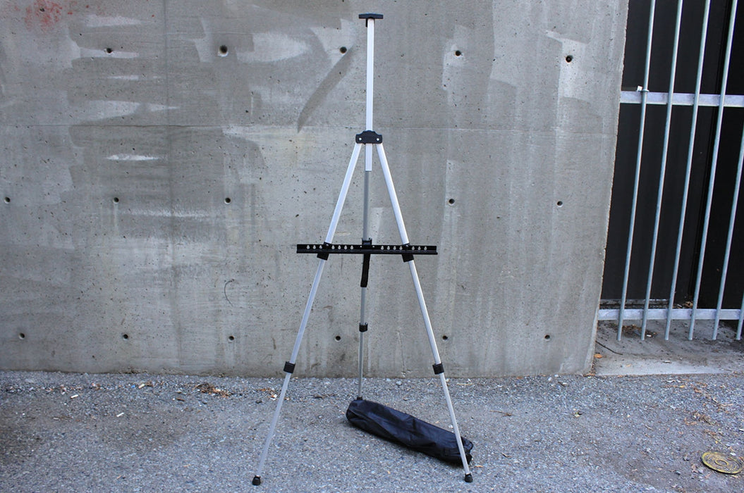 Extended folding aluminum easel stand with transportation bag on a grey concrete background.